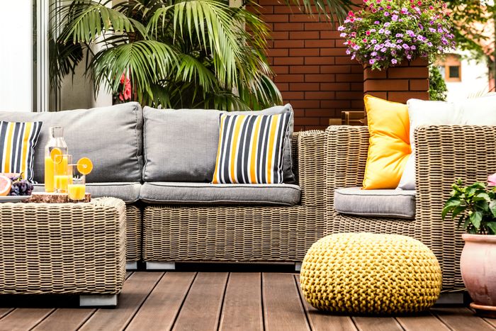 A set of wicker patio furniture covered in grey cushions and yellow pillows, with a red brick back wall.